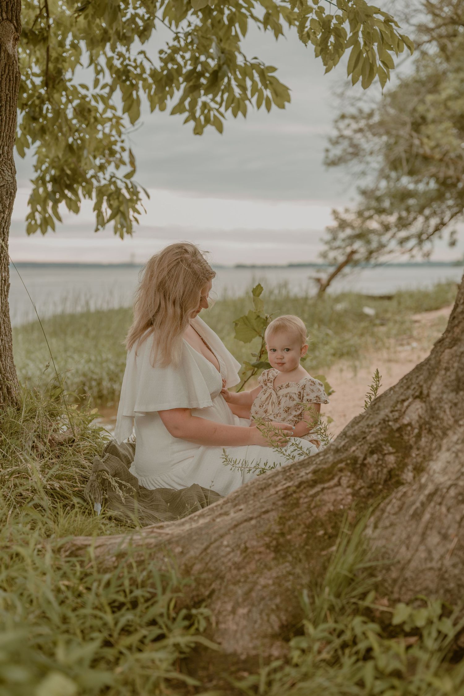A mother and toddler sit together under trees near a lake shore, sharing a peaceful outdoor moment.