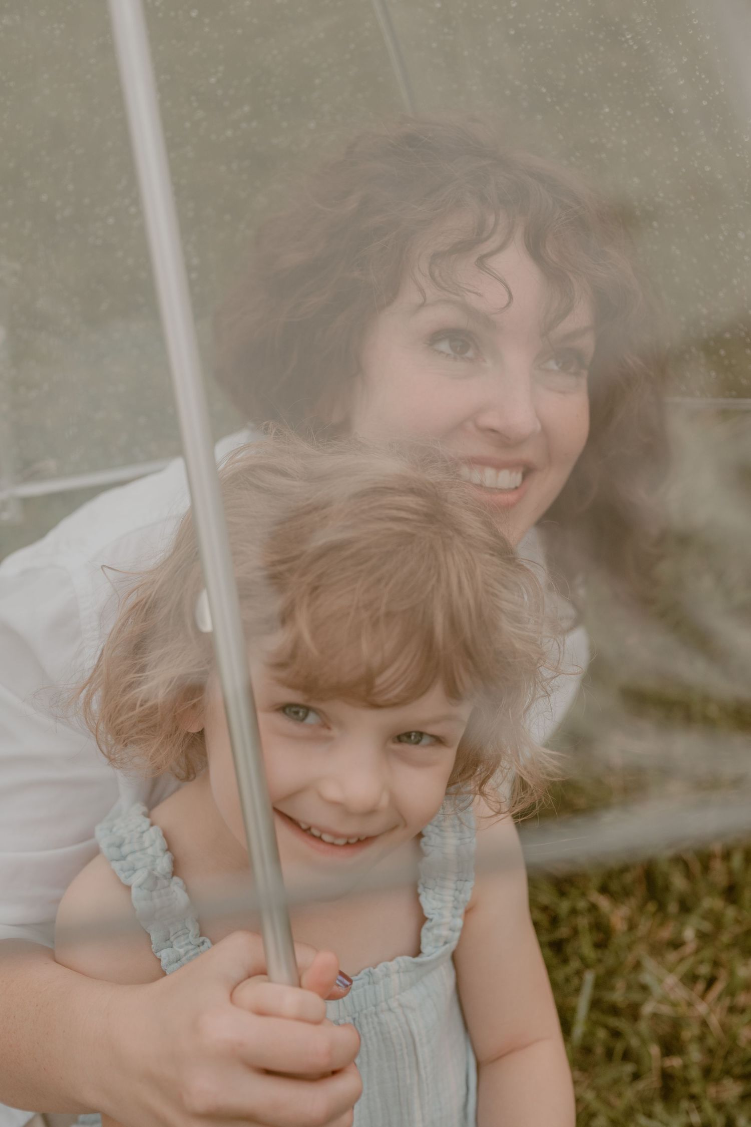A smiling mother and young daughter share a clear umbrella outdoors, both looking happy in casual summer clothing.
