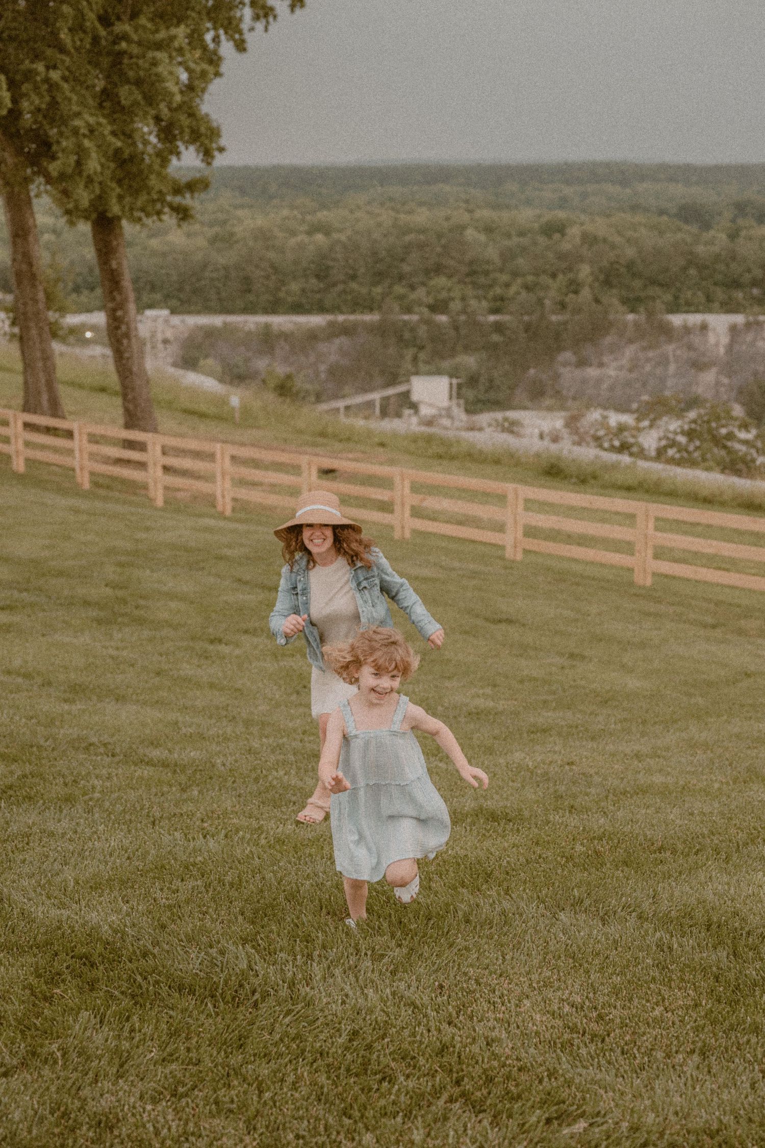 A woman in a denim jacket and hat chases a young girl in a blue dress across a green lawn with a wooden fence.