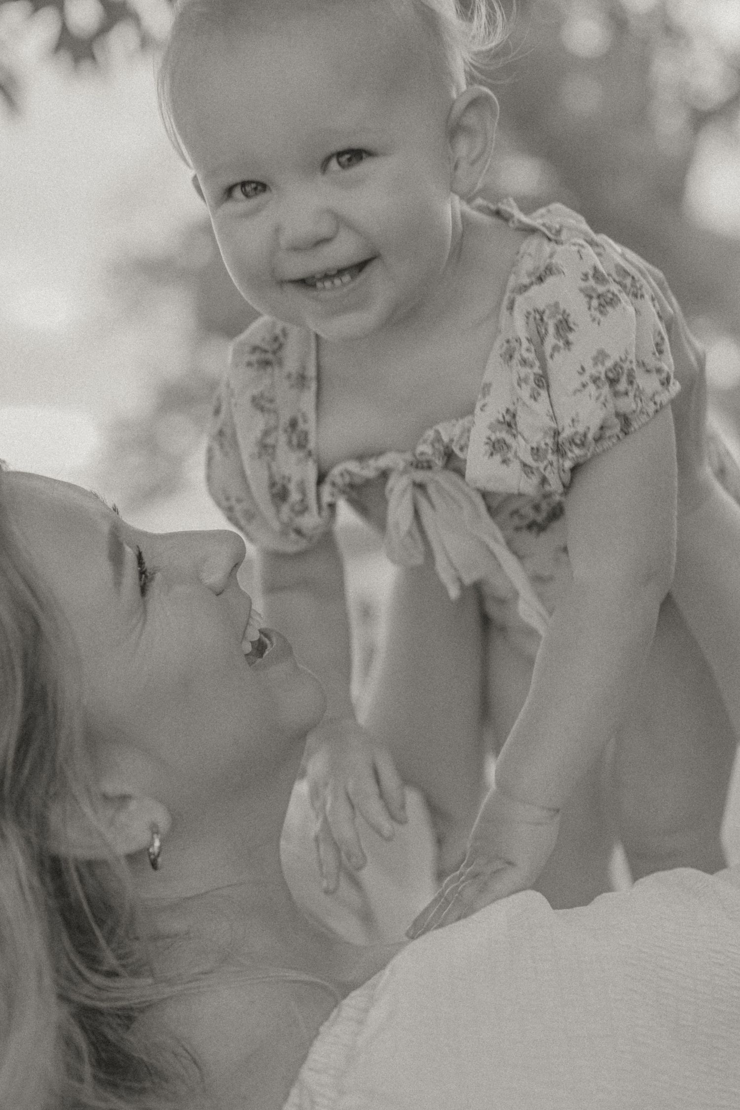 Black and white photo of a smiling baby girl in floral dress being lifted by her mother outdoors.