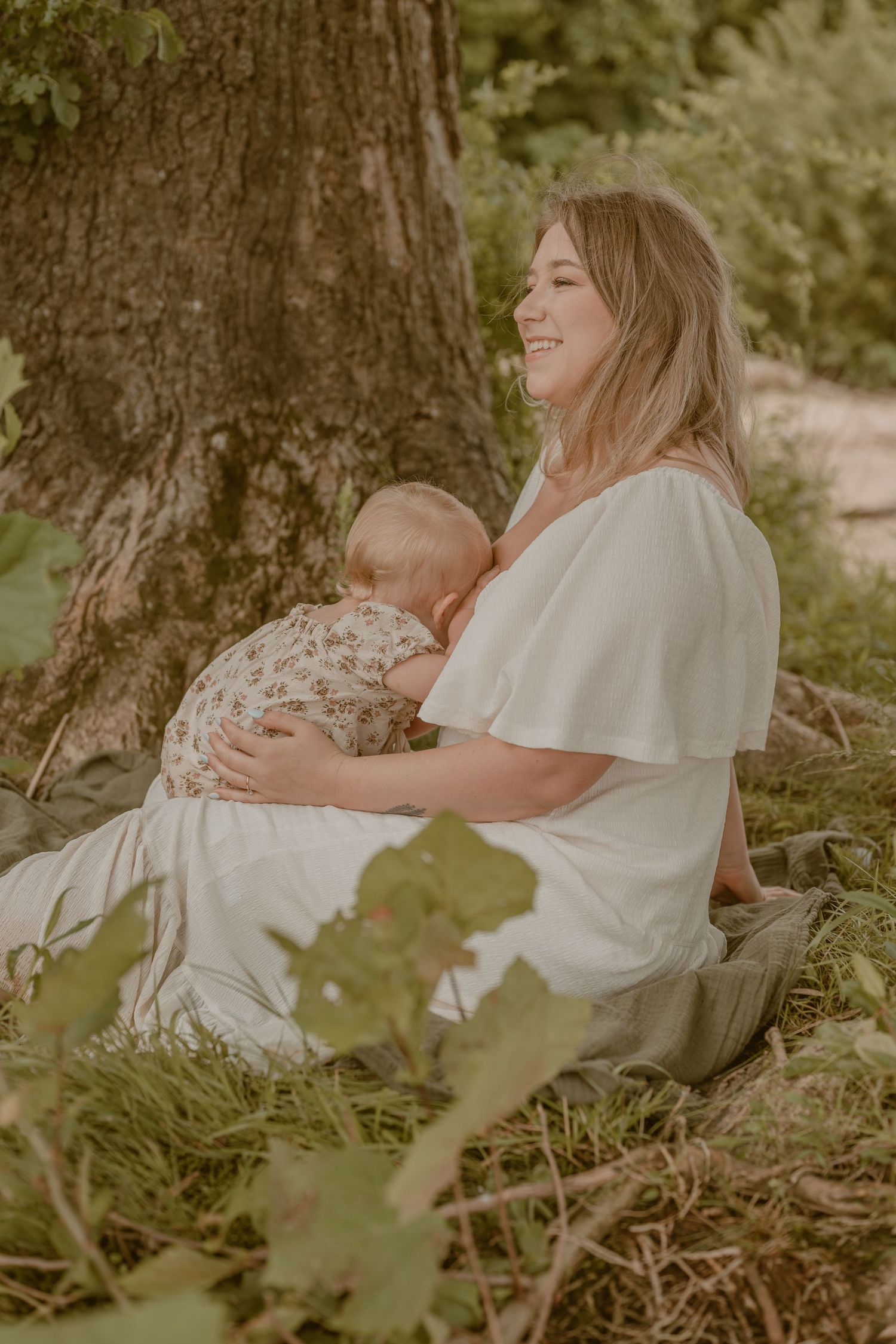 A smiling mother breastfeeding her baby while sitting against a tree in a lush green outdoor setting.