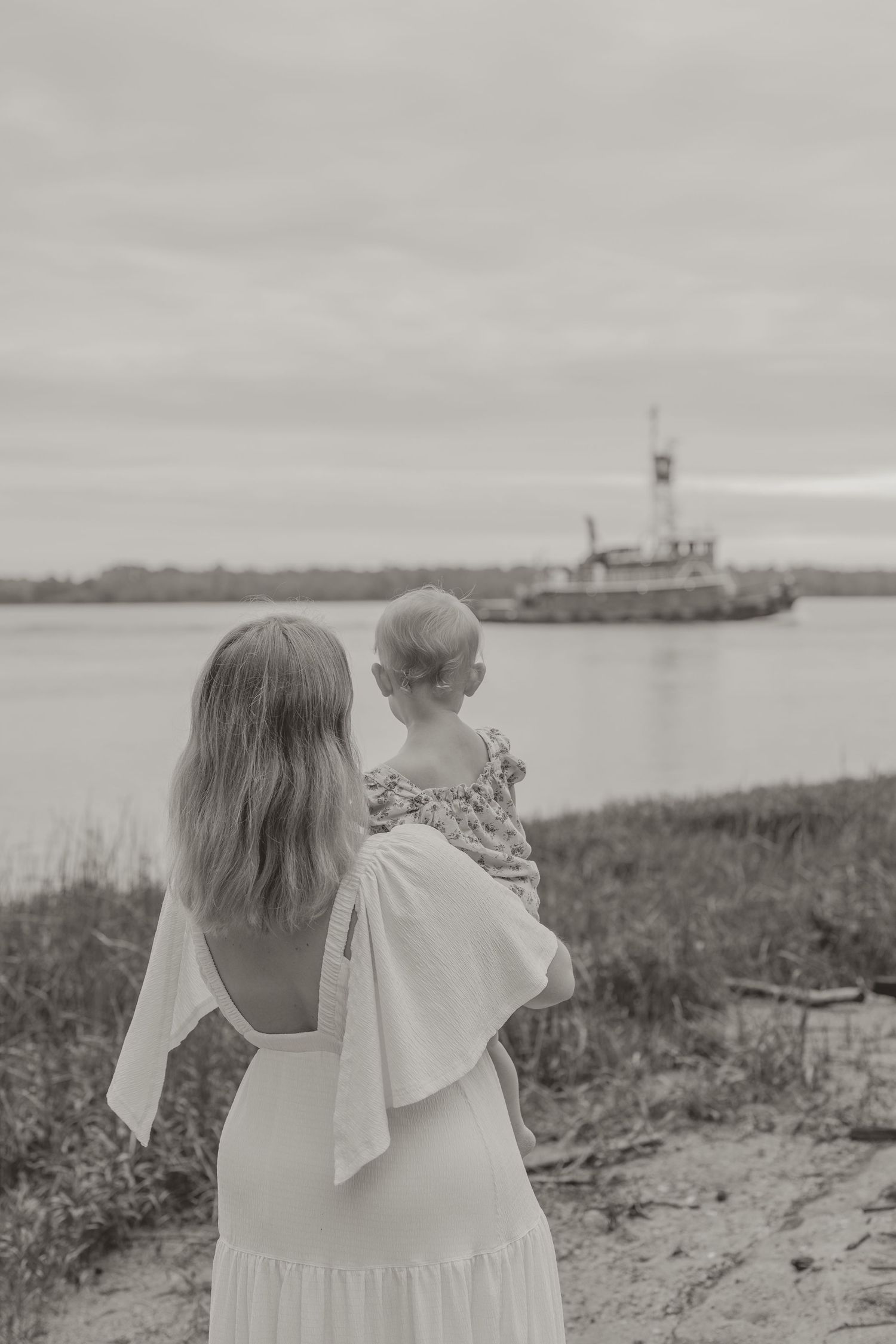 Black and white photo of a mother in white dress holding her baby, gazing at a boat on the water.