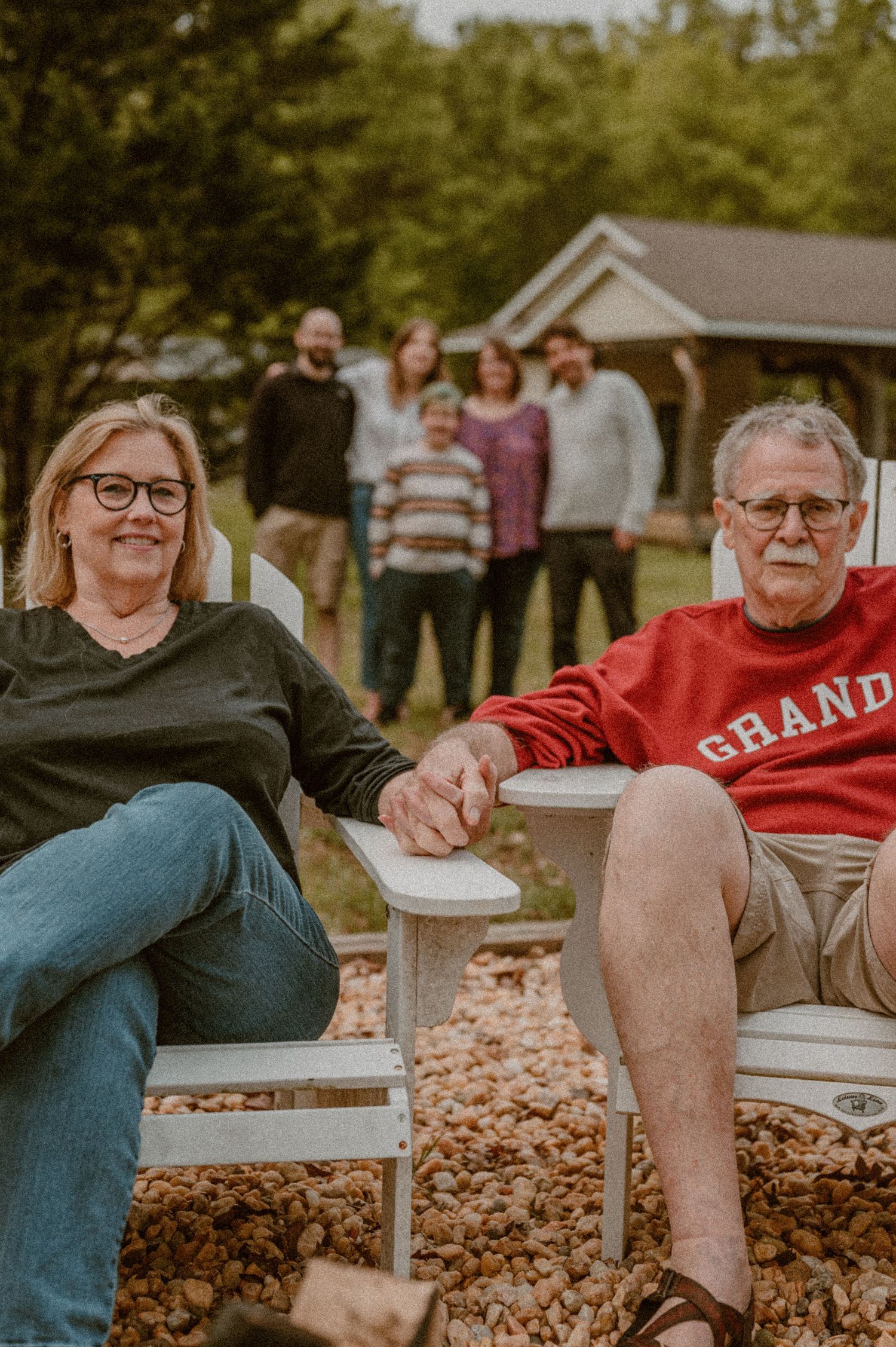 grandparents sit together on outdoor furniture while family pose in the background near a gazebo on a sunny day. photo by Samantha Campbell