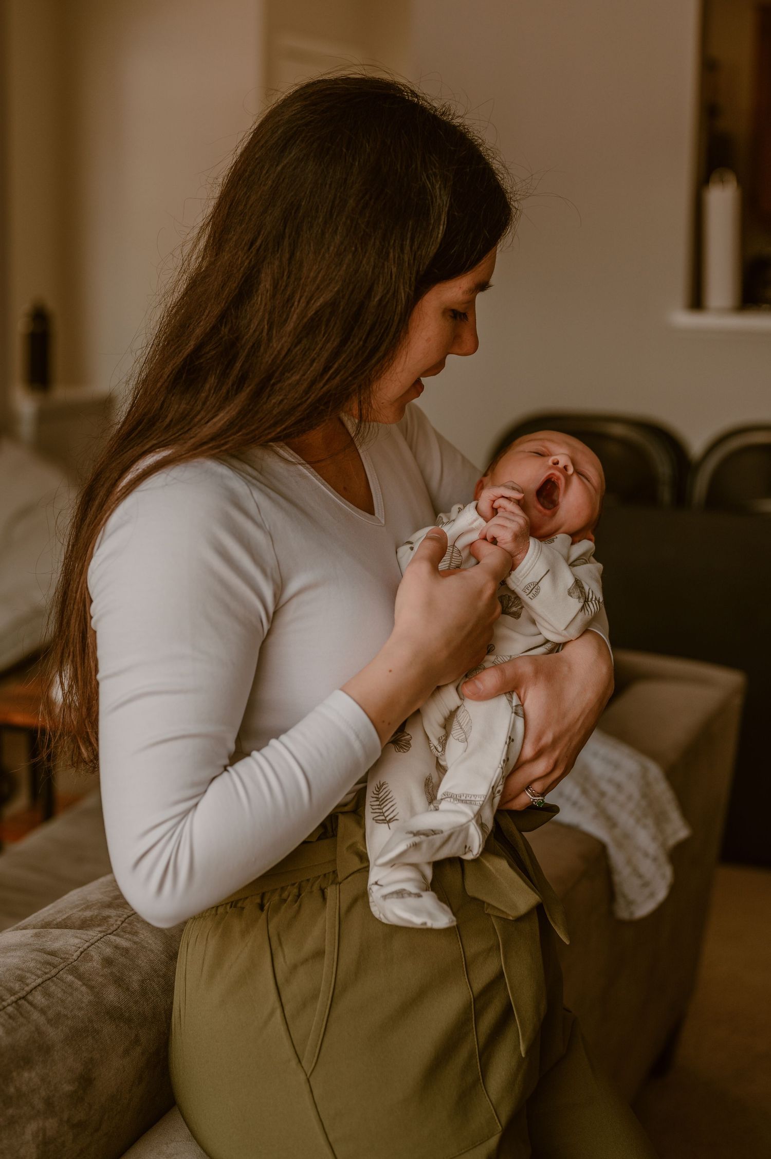 Mother in white holding yawning newborn baby in white outfit while standing against sofa. photo by Samantha Campbell imagery
