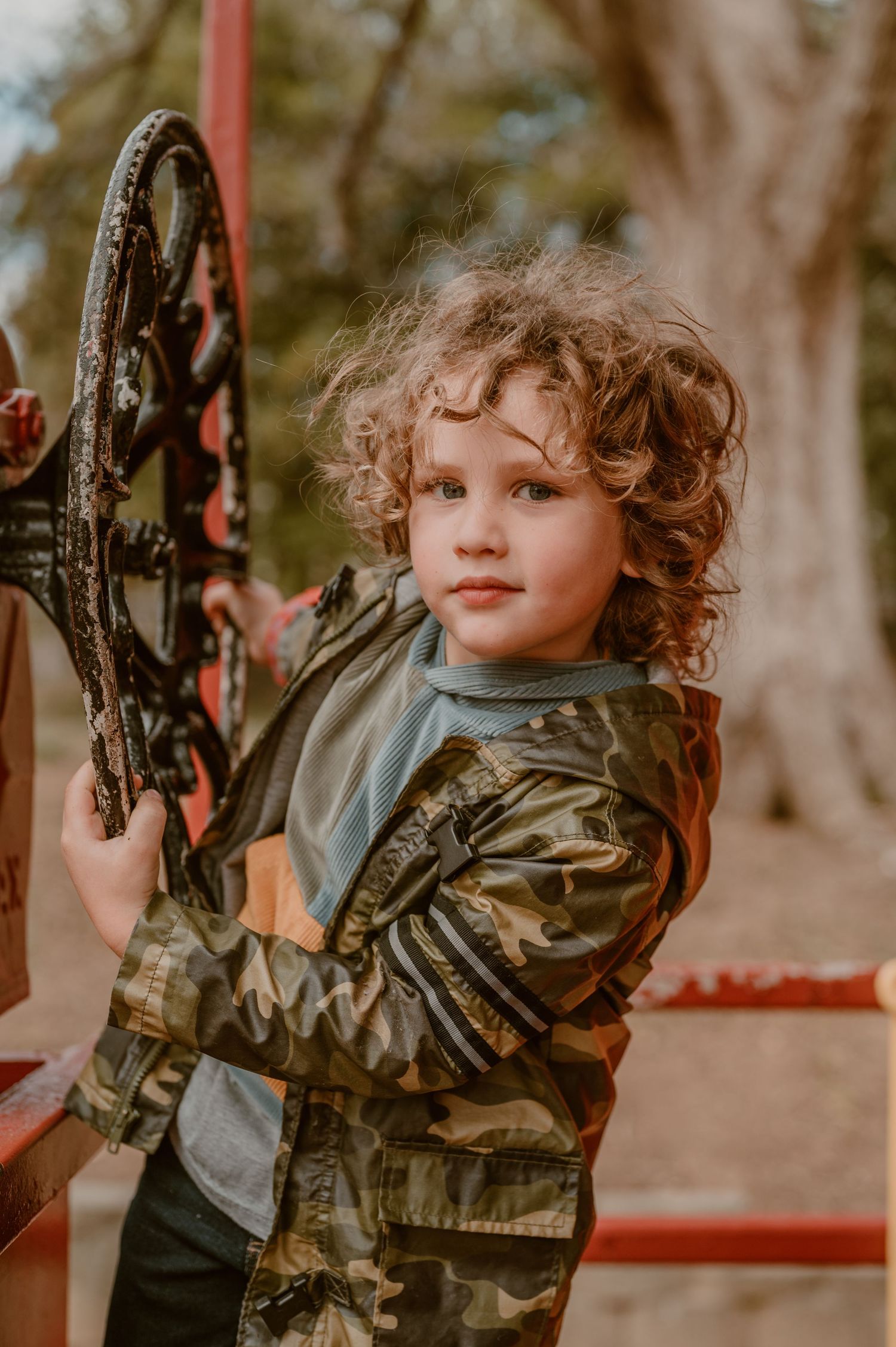 Young child wearing camouflage jacket standing near outdoor playground equipment in natural setting.
