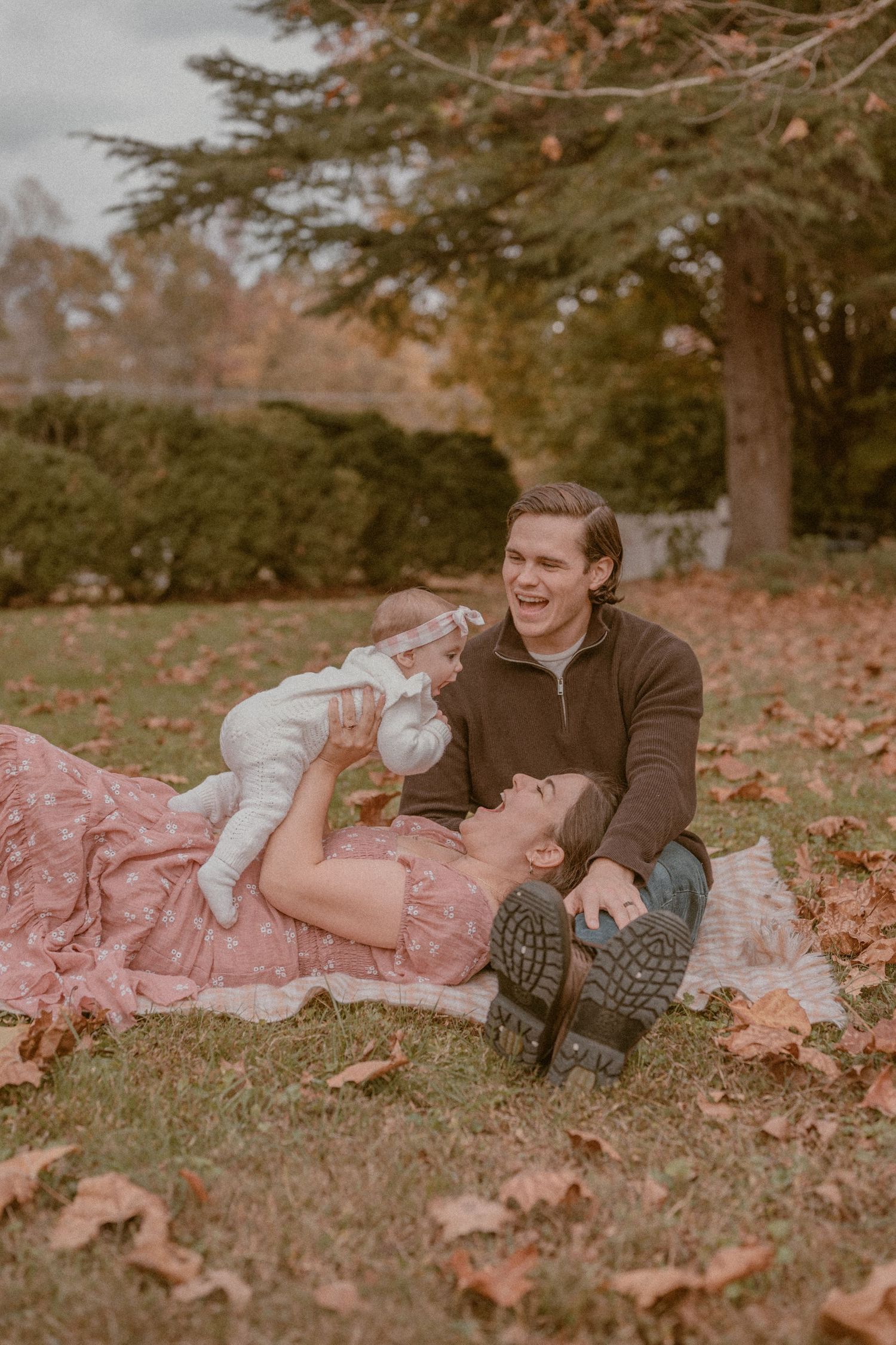 A happy family enjoys an autumn picnic, with a laughing dad and mom holding a baby in a white outfit on fallen leaves.