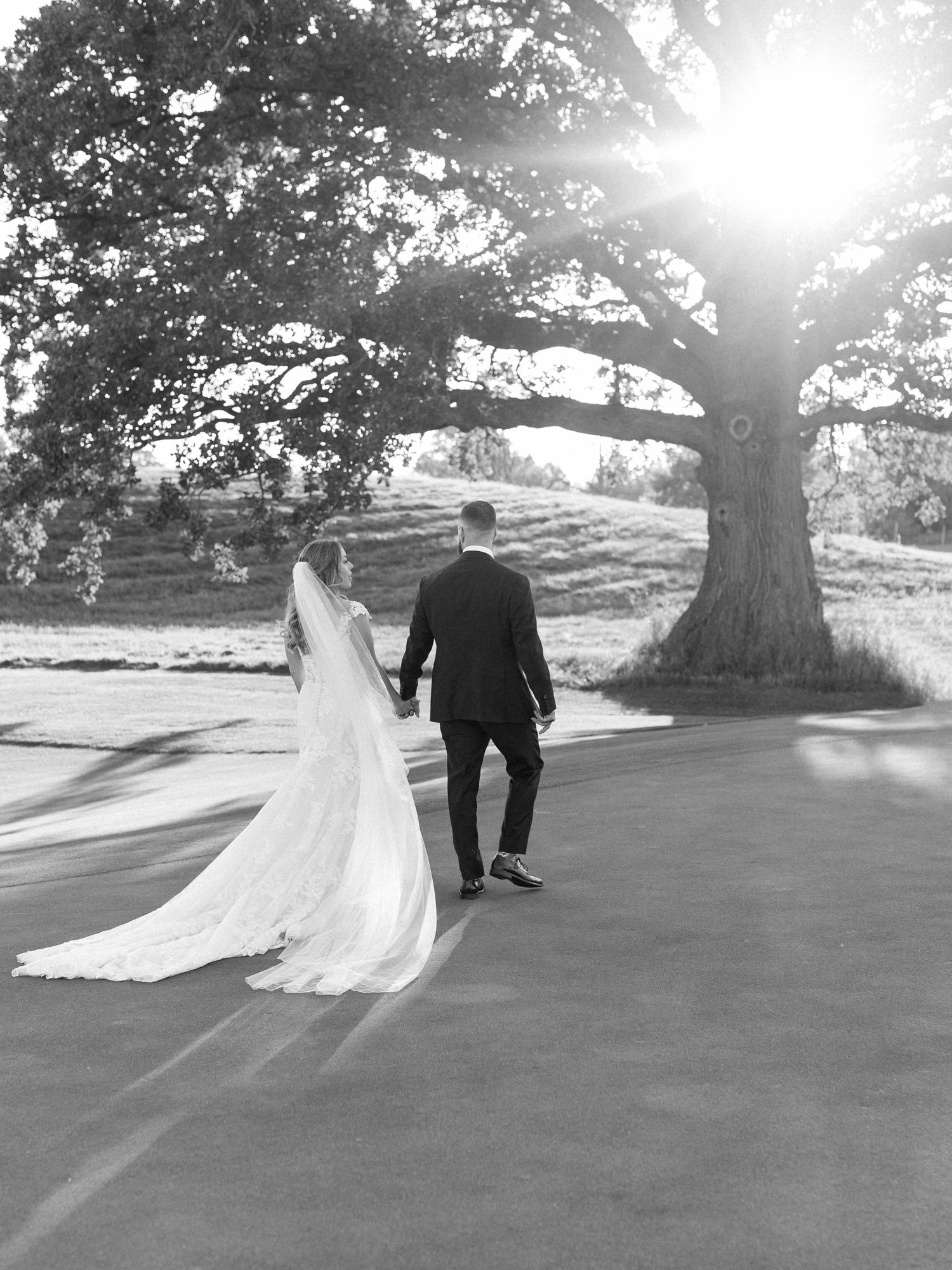 A groom leads his bride to a large willow tree, her dress flowing behind her at Piper's Heath Golf Club.