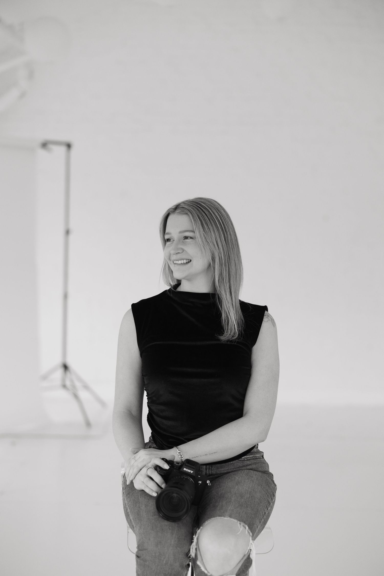 Black and white professional studio photo of someone in a black sleeveless top and jeans sitting casually against white backdrop.