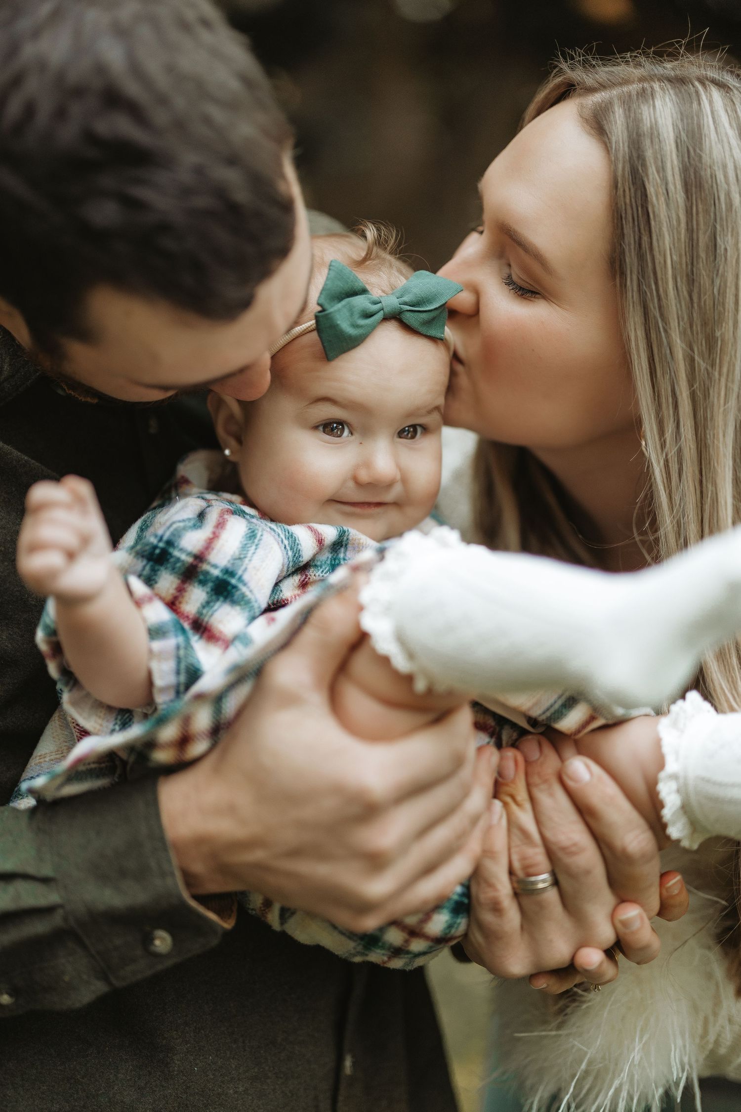 Parents kissing baby with green bow headband in intimate family portrait.