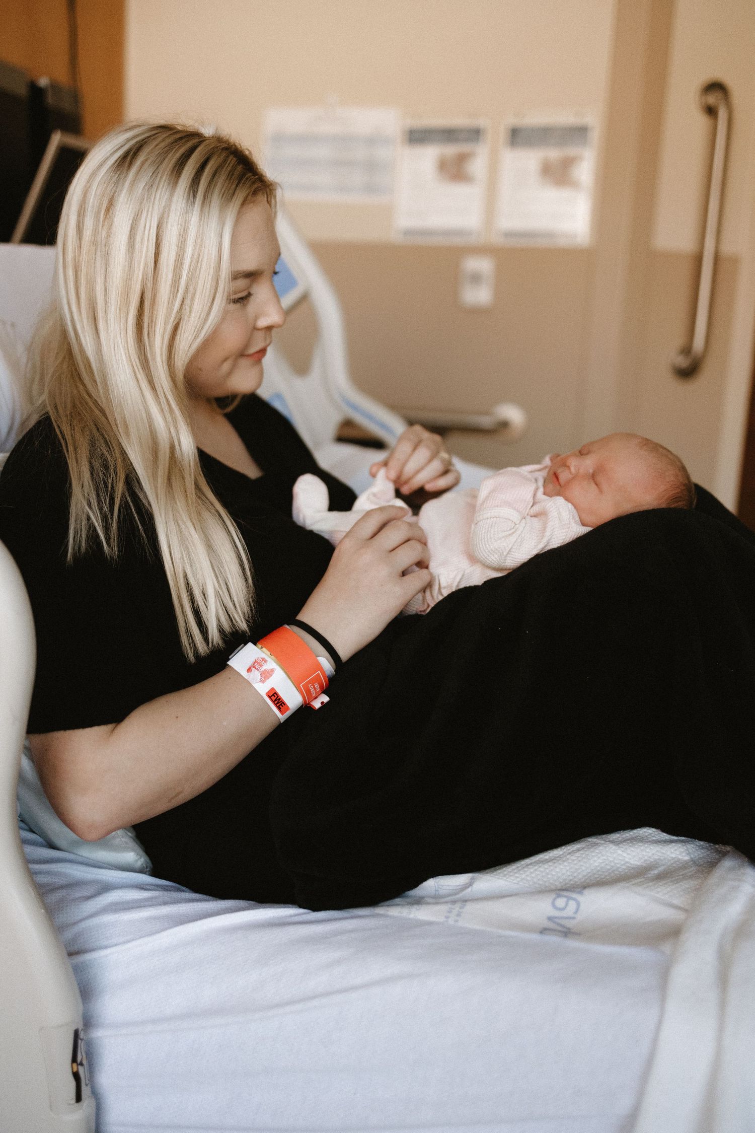 A woman with blonde hair wearing a hospital bracelet cradles her newborn baby in a hospital bed.