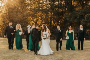 Wedding party poses together in formal attire with bridesmaids in emerald green dresses at golden hour.