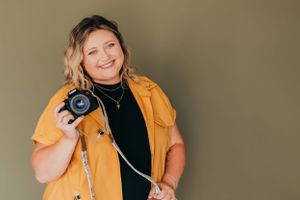 Person in yellow jacket holding camera against neutral background with joyful expression.
