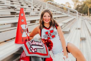 Cheerleader in uniform holding red megaphone while posing on stadium bleachers at sunset.