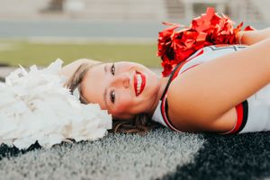 Cheerleader with red pom poms laying on turf field during photo session.