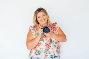 Person in floral dress holding camera against white background with cheerful expression.
