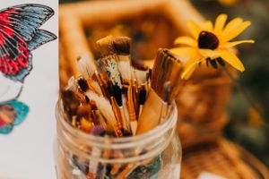 Paint brushes in a glass jar next to butterfly artwork and yellow flowers.