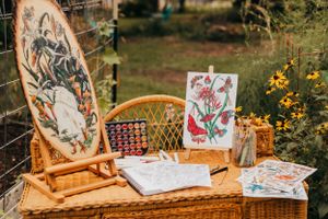 Art supplies and paintings displayed on wicker furniture in a garden setting.