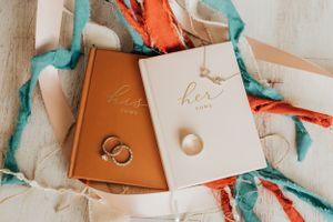 Wedding vow books and rings arranged with coral and turquoise ribbons on white fabric.