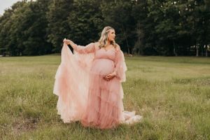 Maternity photo of person in flowing pink gown standing in sunlit field.