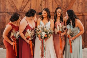 Wedding party in mix-and-match coral and sage dresses holding bouquets by rustic barn door.