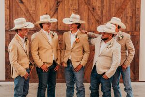 Groomsmen in tan suits and cowboy hats posing against wooden barn door.