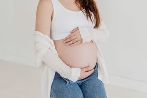 Pregnant person in white tank top and jeans cradles their baby bump while wearing a cozy cream cardigan.