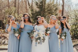 Bridesmaids in dusty blue dresses stand together holding white and purple floral bouquets at an outdoor wedding.