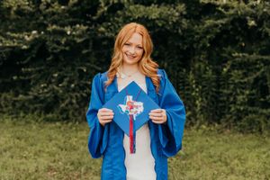 A graduate in blue graduation gown and white dress holds a diploma in a sunny outdoor setting with green foliage.