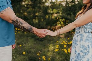 A couple holds hands in a sunlit garden, the man's tattooed arm reaching toward a woman in a floral dress.