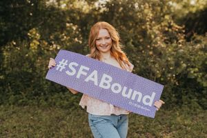 A person holds up a glittery purple sign reading #SFABound in an outdoor setting with green foliage in the background.