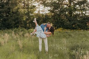 Couple sharing a romantic kiss in a lush green meadow, man lifting woman in floral dress at golden hour.
