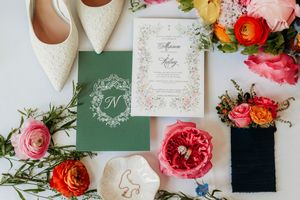 Wedding details flatlay with ivory shoes, floral wedding invitations in green and white, and pink and coral roses.