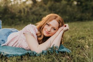 A person in a pink floral dress lies on their side on a blue blanket in green grass during golden hour.