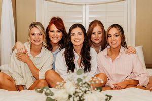 Five women wearing soft neutral robes gather together on a couch with white florals in the foreground.