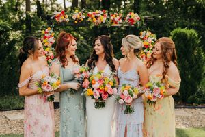 A group wearing pastel floral dresses poses together amid colorful flowers in a garden setting.