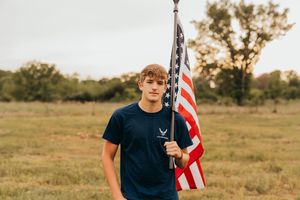 Someone in an Air Force t-shirt stands in a grassy field at sunset while proudly holding an American flag.