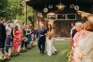 Newlyweds walk down outdoor aisle decorated with lanterns and floral arrangements at rustic barn wedding ceremony.