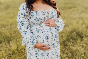 A pregnant figure stands in a sunlit meadow wearing a flowy white and blue floral off-shoulder dress.