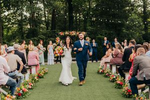 Guests watch a couple walk down a floral-lined outdoor ceremony aisle under string lights in a wooded garden setting.