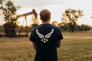 Person wearing US Air Force t-shirt with wings logo stands facing oil pump jack in rural field at sunset.