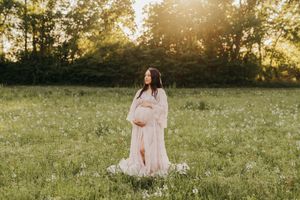 A maternity photo in a dreamy sunlit field shows an expectant mother in a flowing pink dress at golden hour.