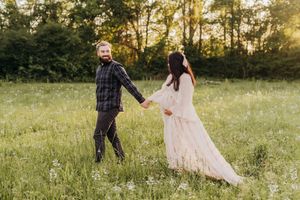 A couple holds hands while walking through a sunlit meadow, with the person in white wearing a flowing dress.
