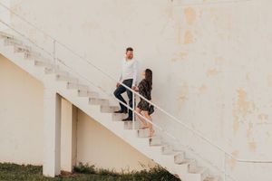 Two people in dark clothing sit together on white concrete stairs against a weathered wall in natural light.