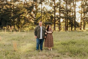 A couple stands together in a sunlit meadow surrounded by tall pine trees during golden hour.