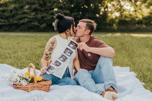 A couple shares a romantic picnic on a white blanket in a sunny park with a wicker basket of food.