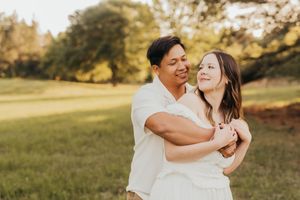 A couple in white outfits embrace while lying in a sunlit grassy meadow during golden hour.