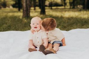Two young children sit together on a white blanket outdoors in a sunlit wooded setting.