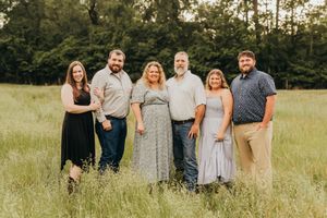 A family group wearing coordinated gray and beige outfits poses together in a sunlit grassy field at golden hour.