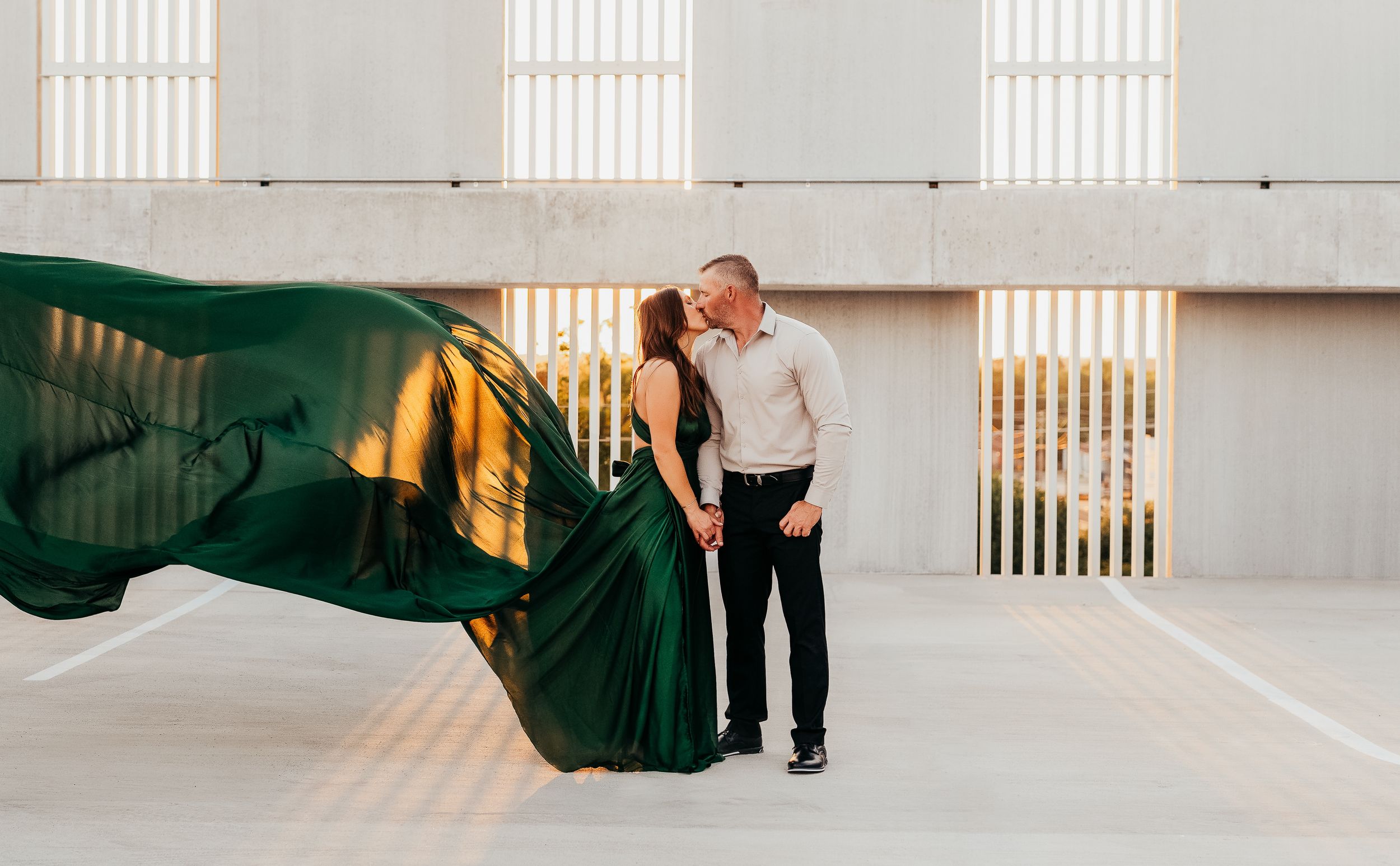 Couple embracing outdoors with green flowing dress billowing in wind against modern concrete architecture backdrop.