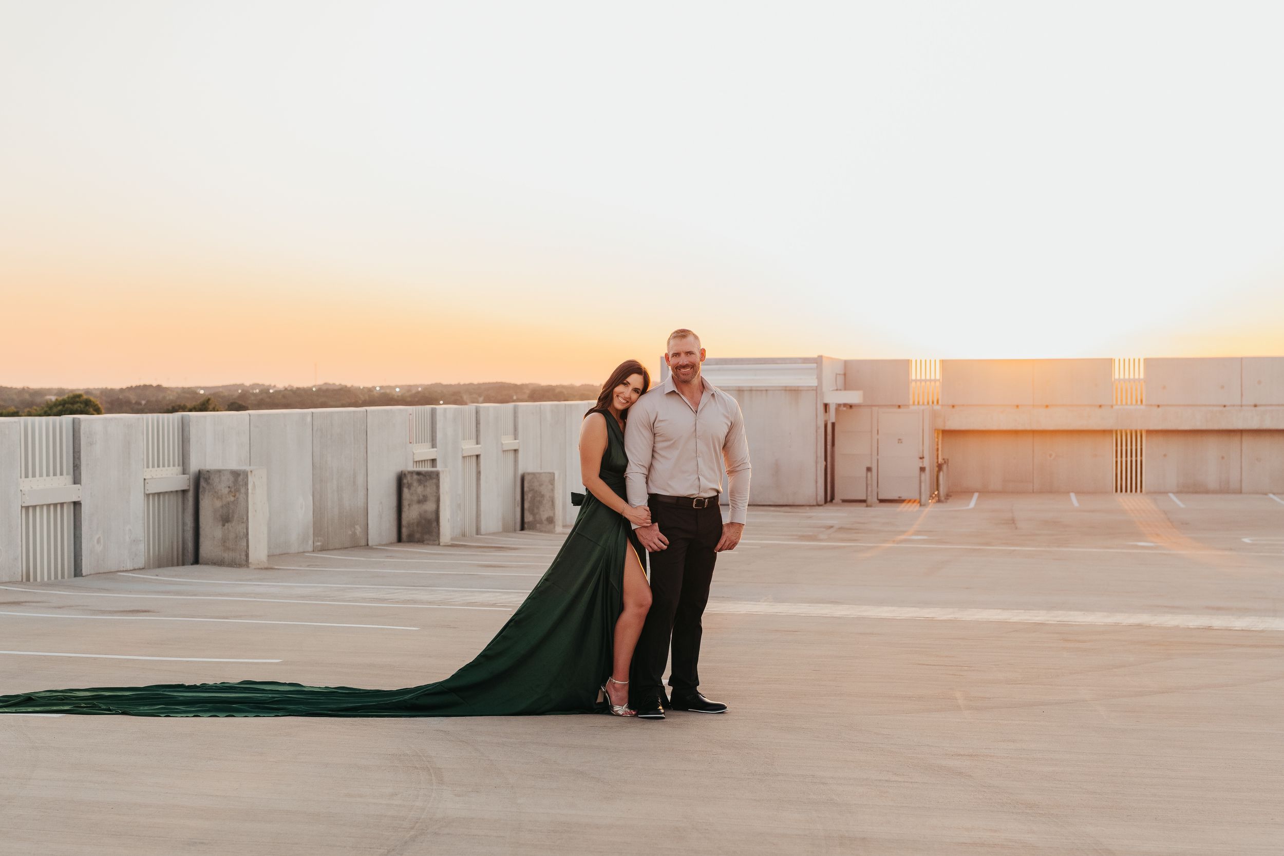 Couple posing at sunset on a rooftop, woman in flowing green gown, man in light shirt, warm golden sky backdrop.